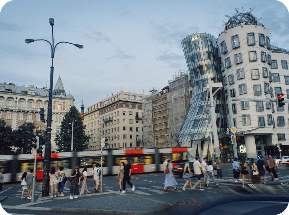 City street with a tram passing by, people walking, and modern buildings, including a uniquely shaped glass structure, under a cloudy sky.