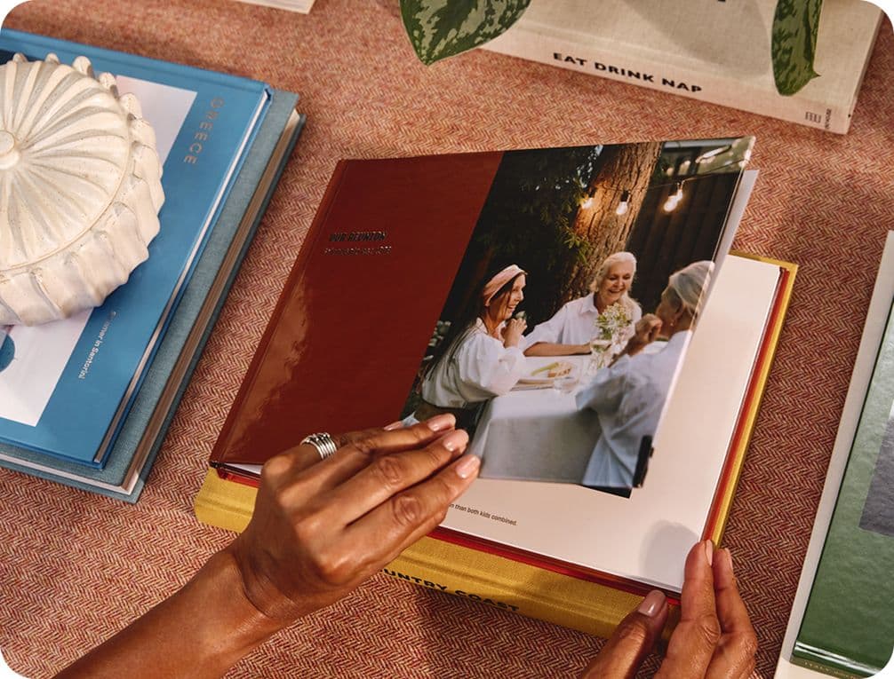 Hands flipping through a photo book showing a group of people dining outdoors. Other books and a decorative item are on the table.