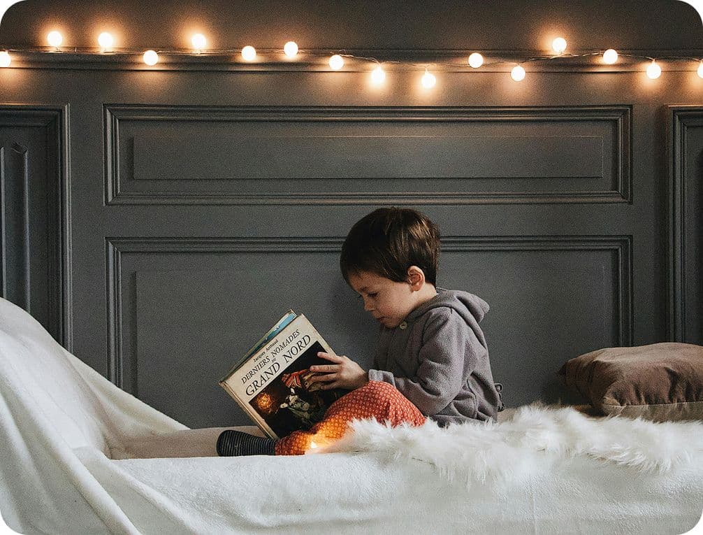 Young child in a cozy room reads a book on a bed with string lights above, creating a warm and inviting atmosphere.