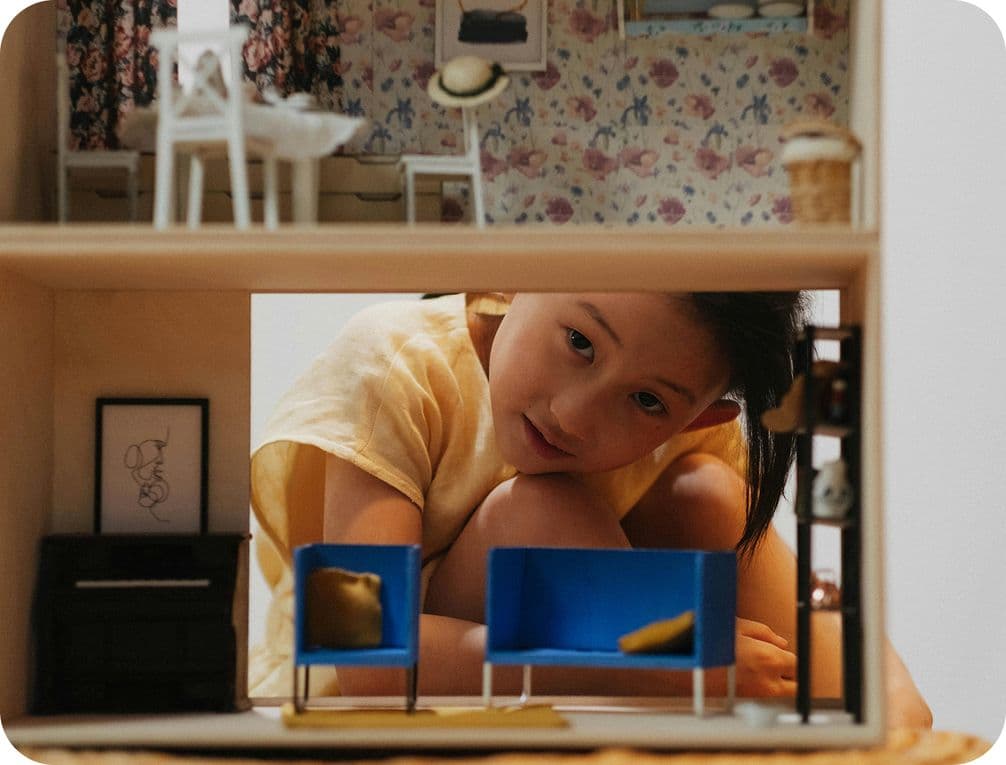 A child in a yellow shirt peers into a dollhouse with blue chairs and floral wallpaper, creating a playful and curious scene.