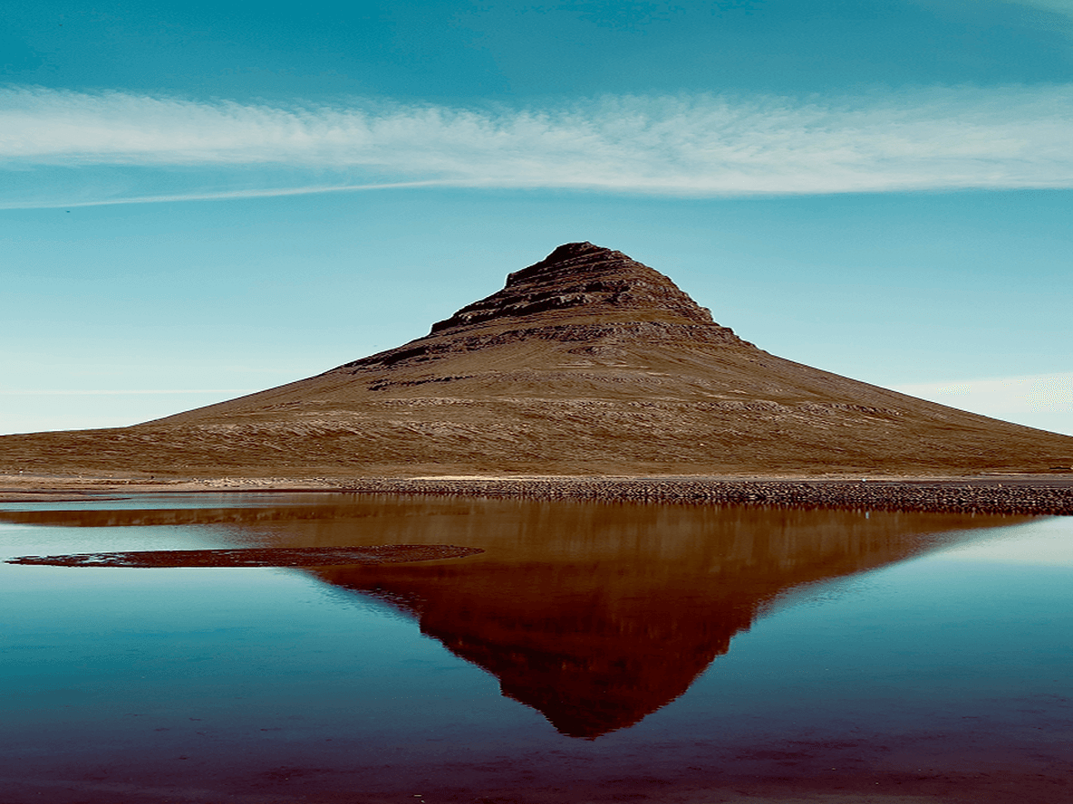 A conical mountain reflected in a calm lake under a clear blue sky, with a thin layer of clouds stretching horizontally.
