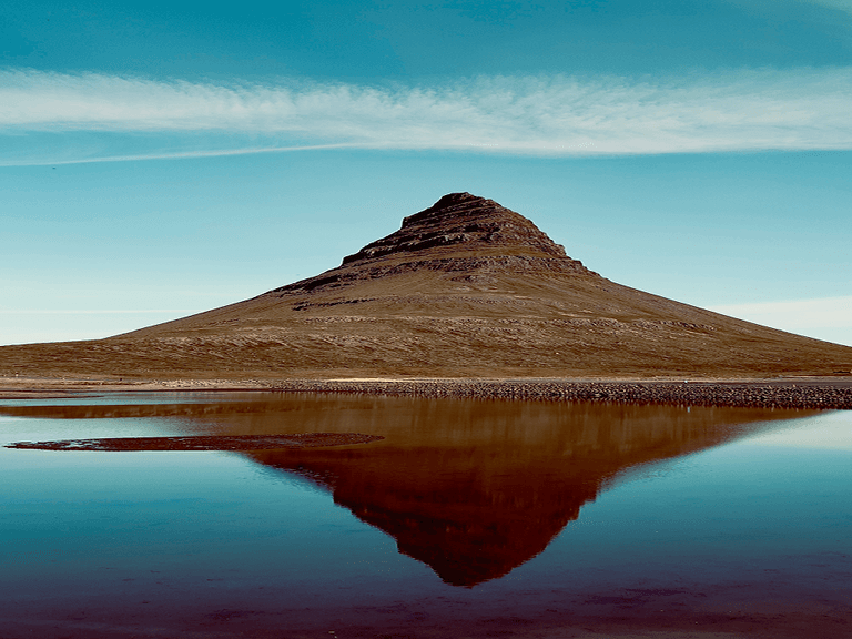 A conical mountain reflected in a calm lake under a clear blue sky, with a thin layer of clouds stretching horizontally.