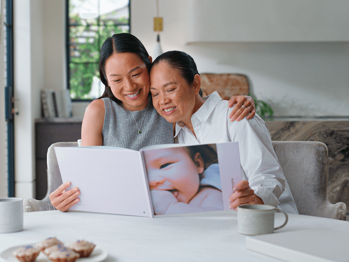 Two women sitting at a table, smiling and looking at a photo album in a bright kitchen