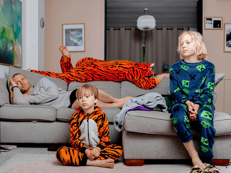 Three people in colorful pajamas relax in a living room; one lies on a couch, while two children sit on the floor and couch.