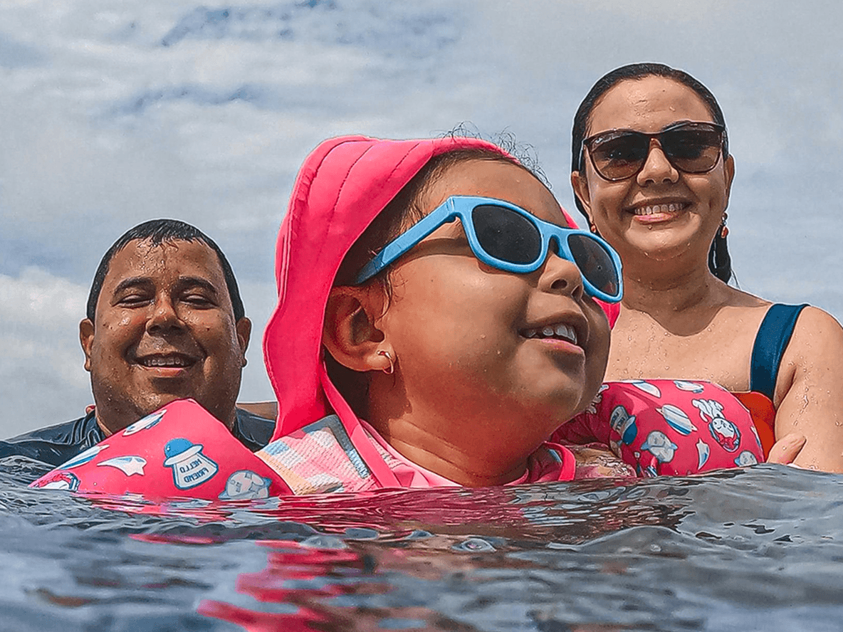 A family of three enjoys swimming in the sea. The child wears pink gear and blue sunglasses, with the parents smiling in the background.