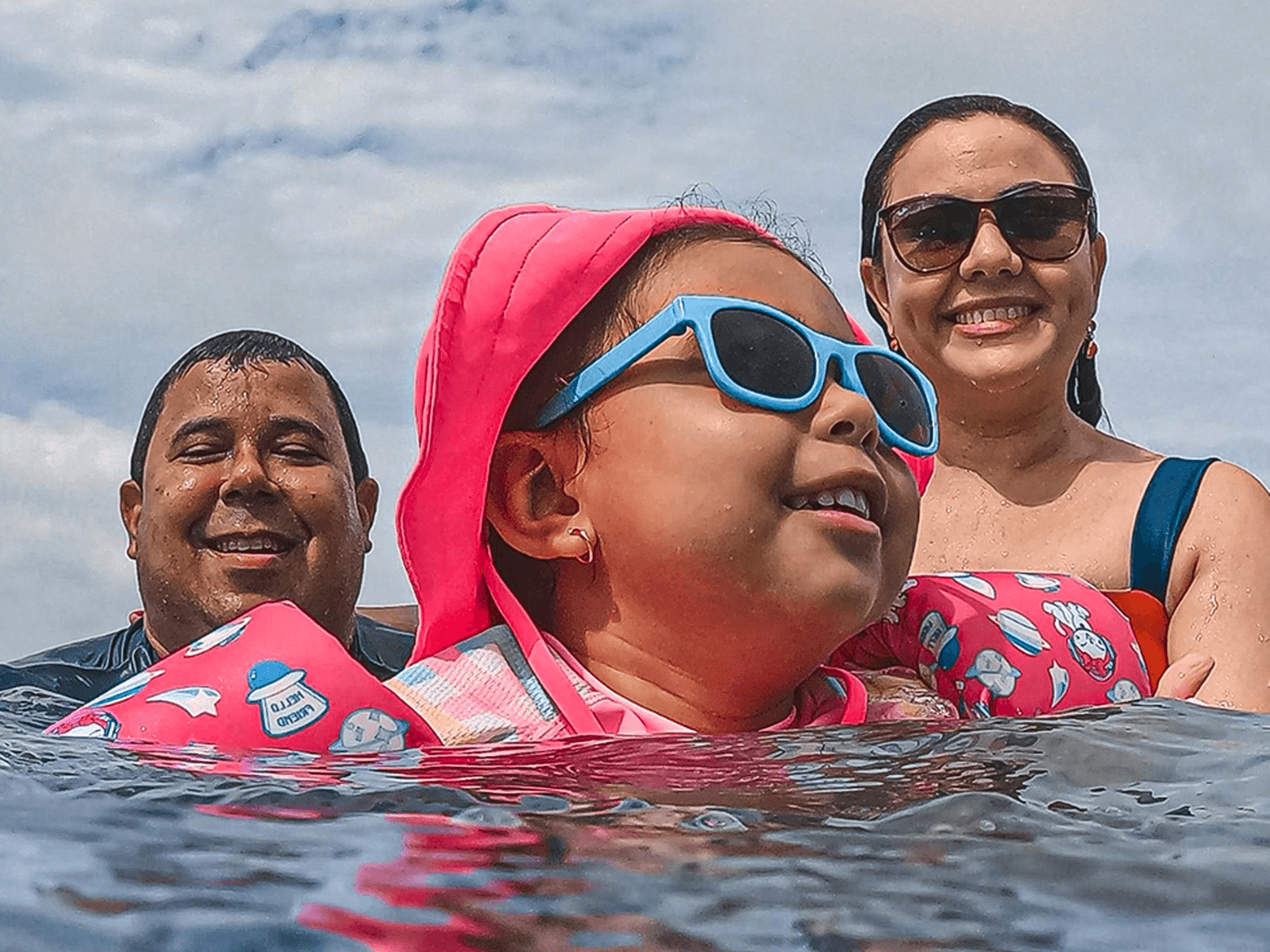 A family of three enjoys swimming in the sea. The child wears pink gear and blue sunglasses, with the parents smiling in the background.