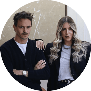 A man and woman stand together indoors, both wearing dark cardigans. The woman has long, wavy hair, and the man has short, styled hair.