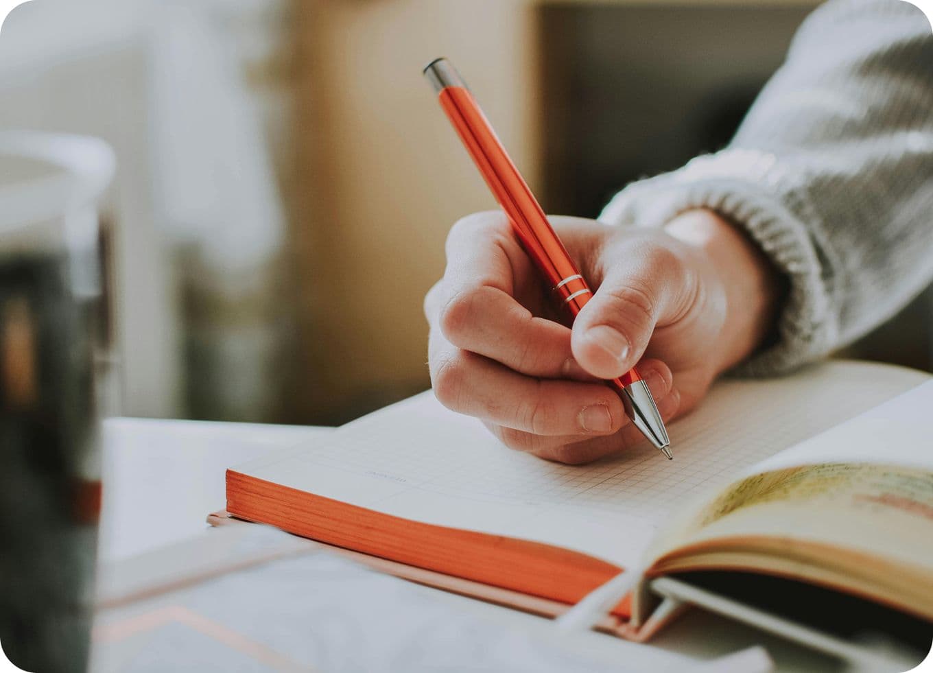 Hand writing with a red pen in an open notebook, with a blurred background and a cup partially visible on the side.
