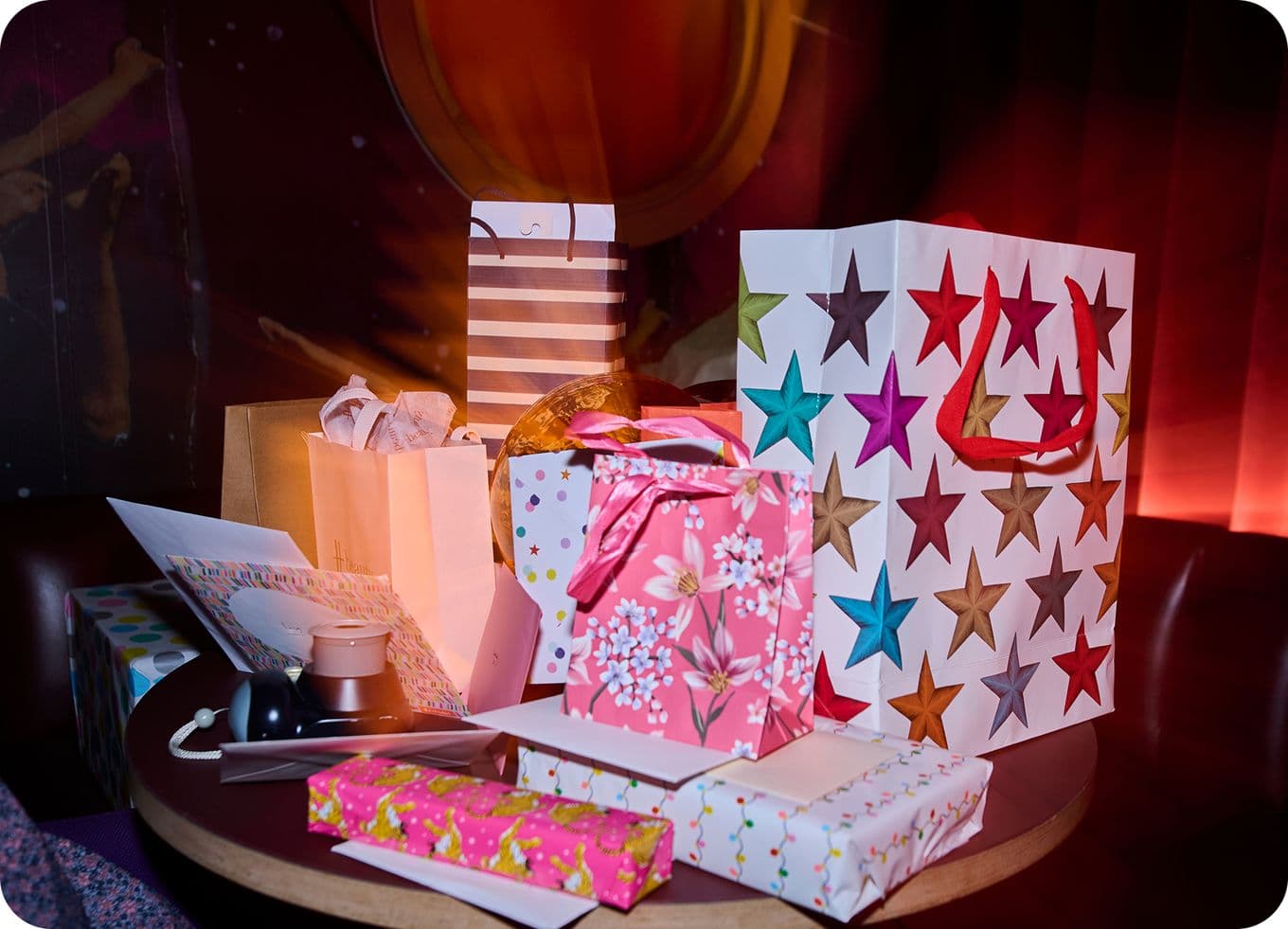 A table filled with colorful gift bags and wrapped presents, featuring star and floral patterns, under warm ambient lighting.
