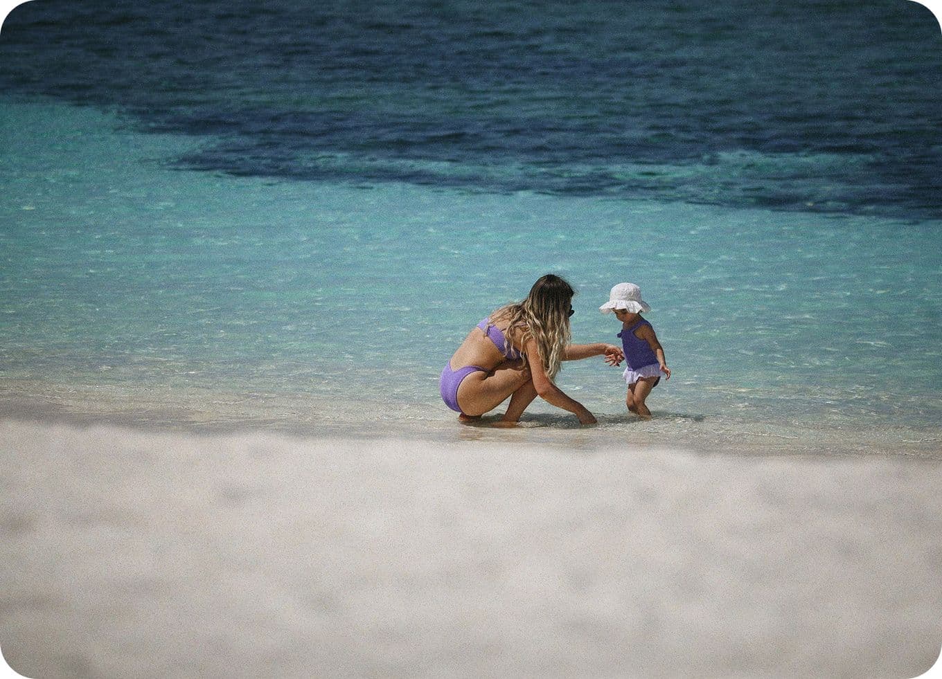 A woman and a child in swimsuits play at the edge of clear turquoise water on a sandy beach.