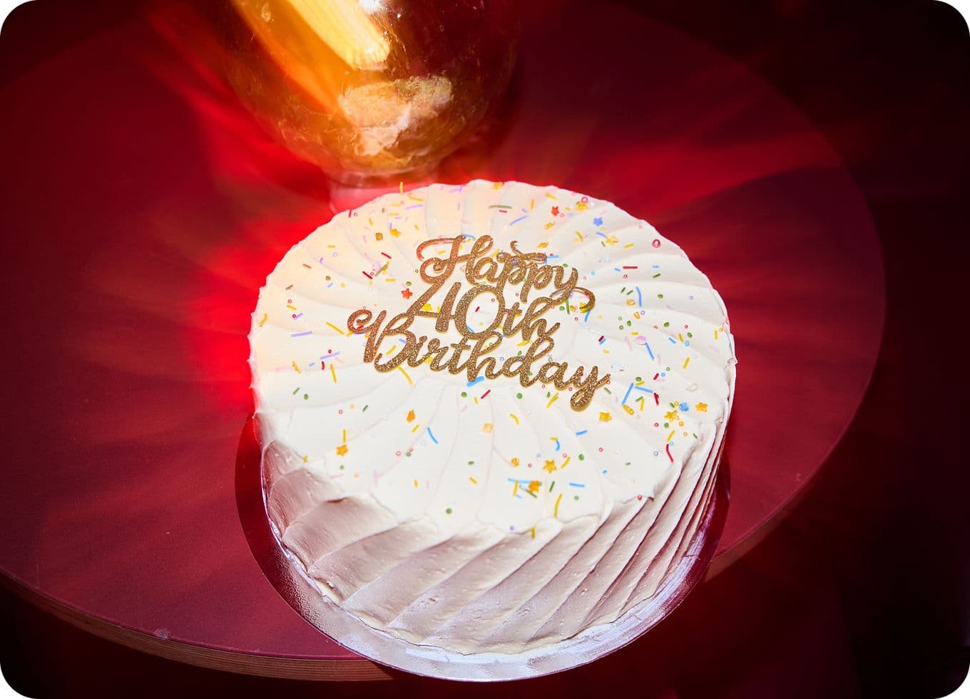 White frosted cake with "Happy 40th Birthday" in gold letters, topped with colorful sprinkles, on a red table.