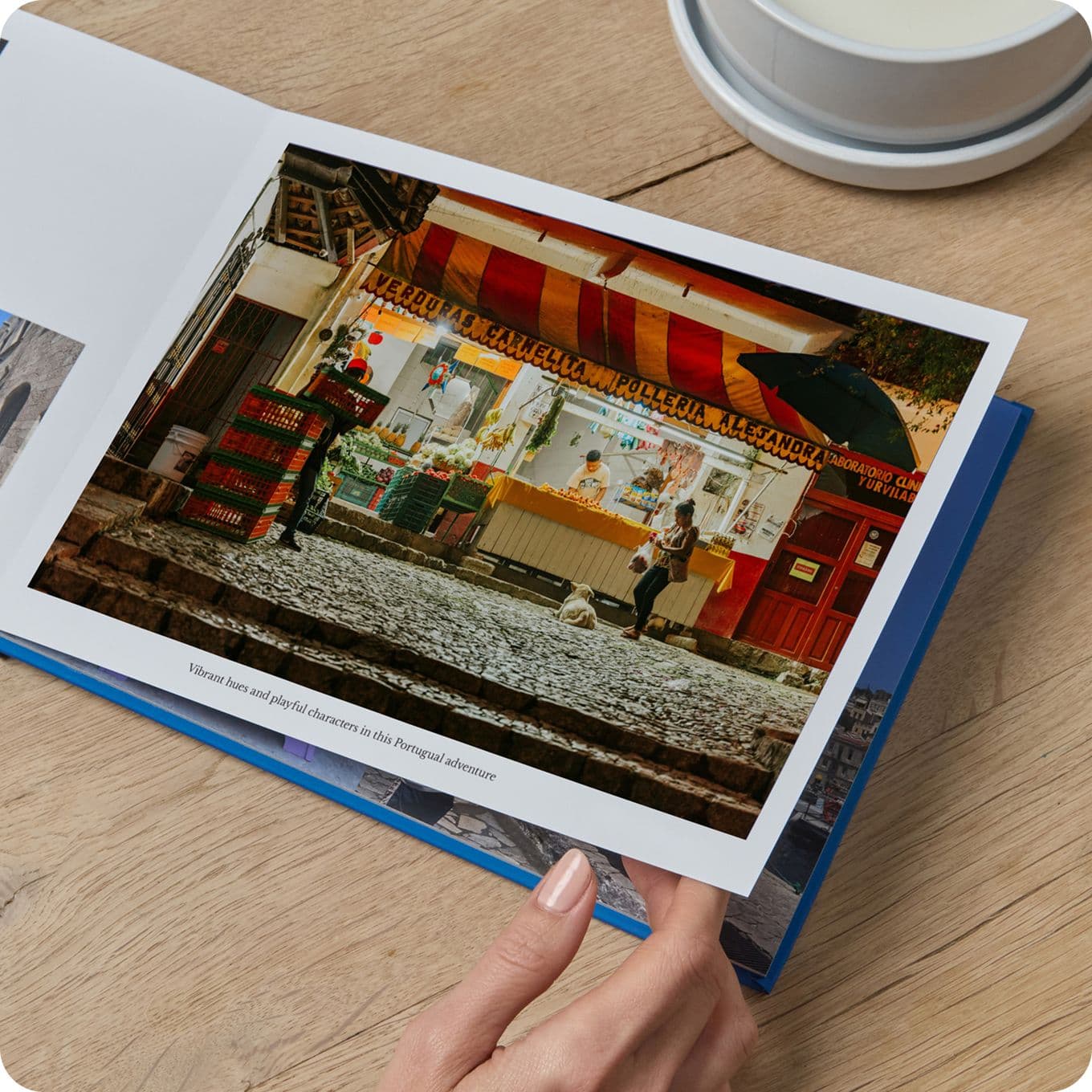 Person holding a photo book open to an image of a vibrant outdoor market with a striped canopy, crates, and a cobblestone path.