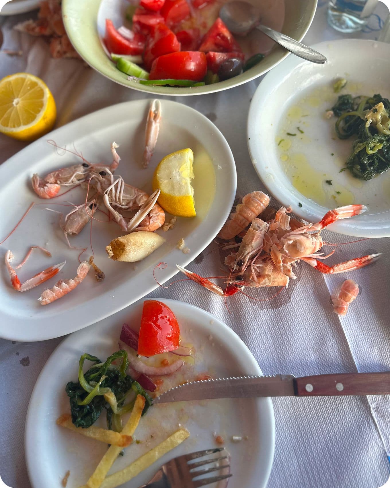Plates with shrimp shells, lemon, salad, and leftover food on a table with a fork and knife.