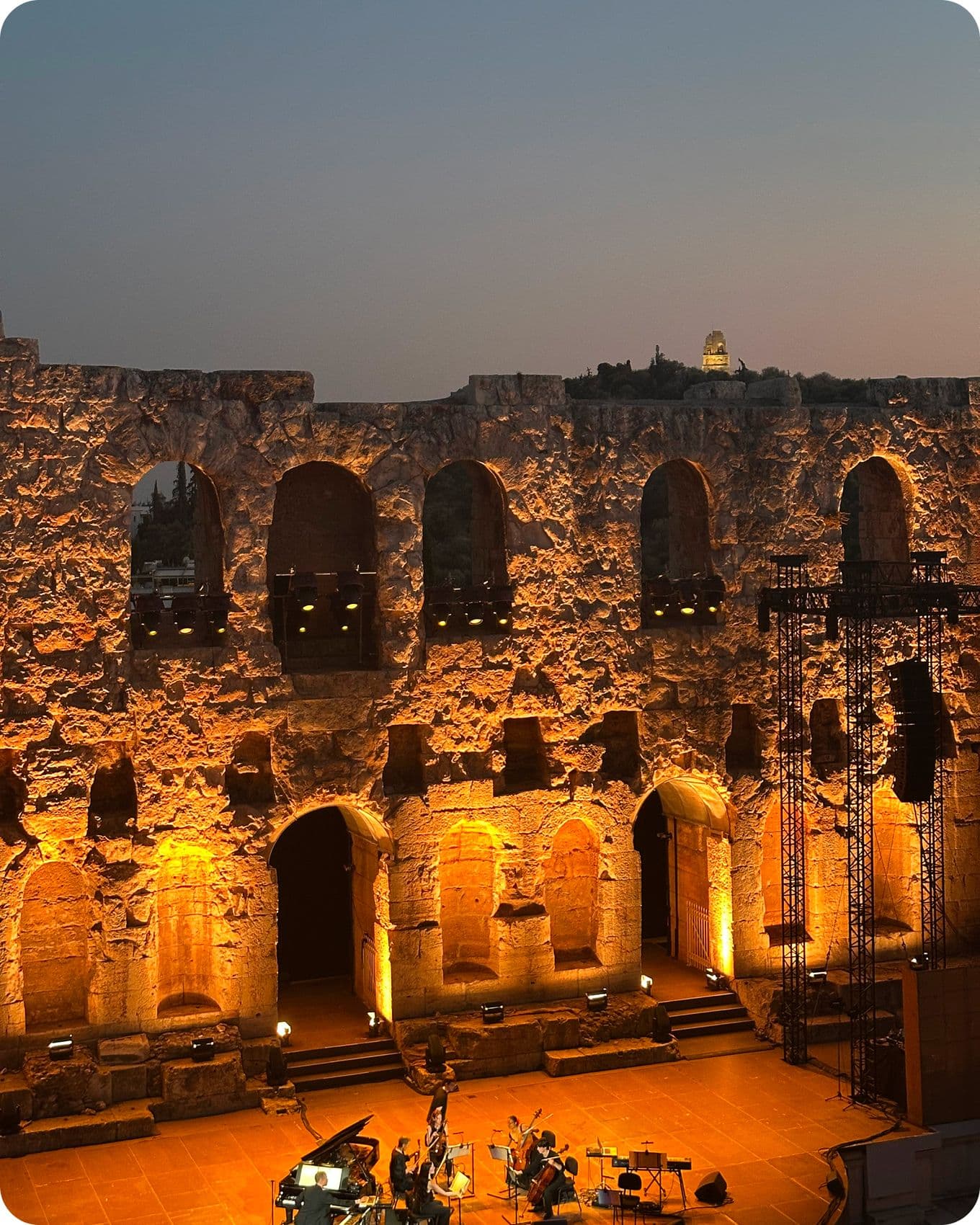 Ancient stone amphitheater illuminated at dusk, with a stage set for a performance and a lit structure visible on a distant hill.