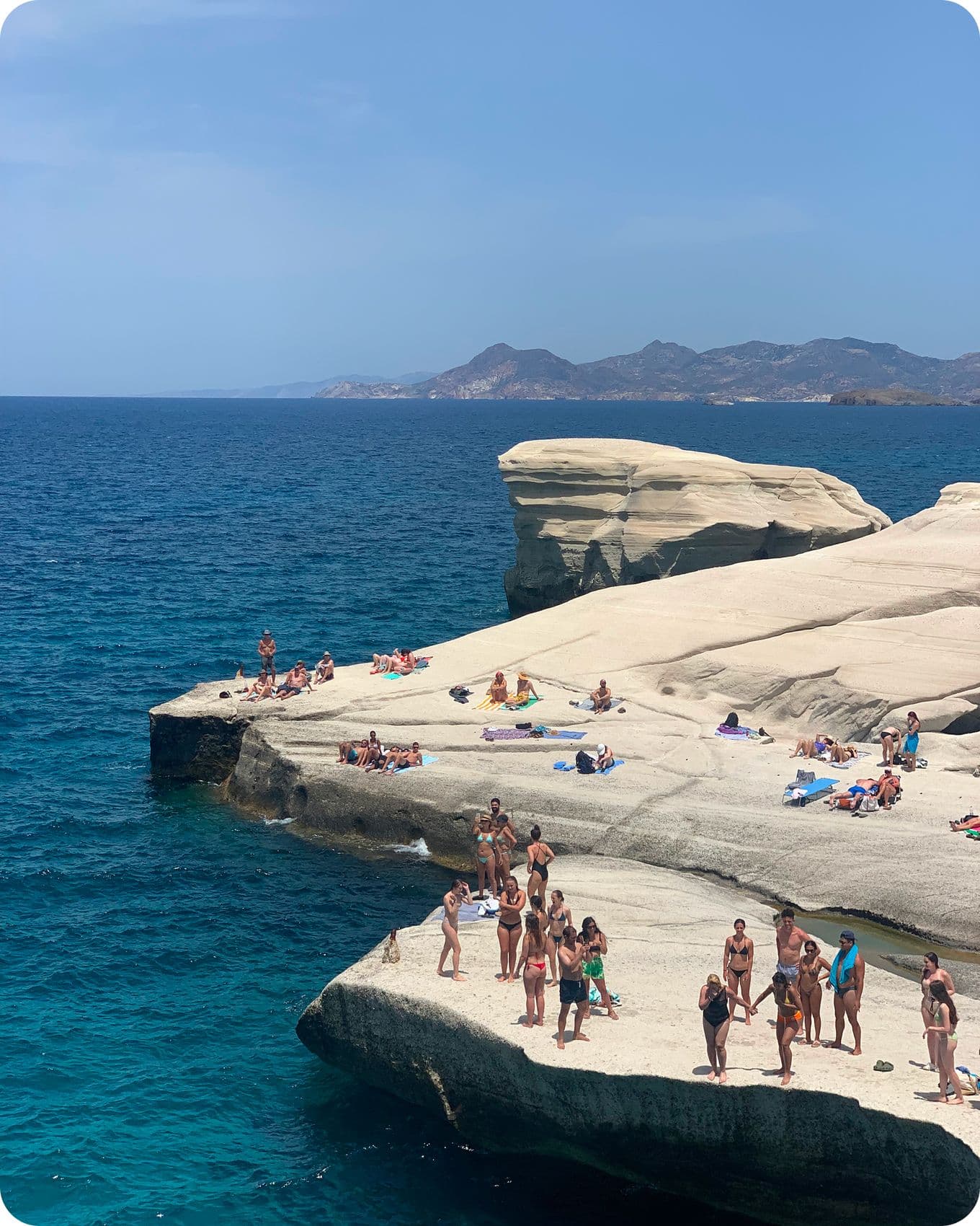 People relax on sunlit, white rocky cliffs by a deep blue sea, with distant hills under a clear sky.