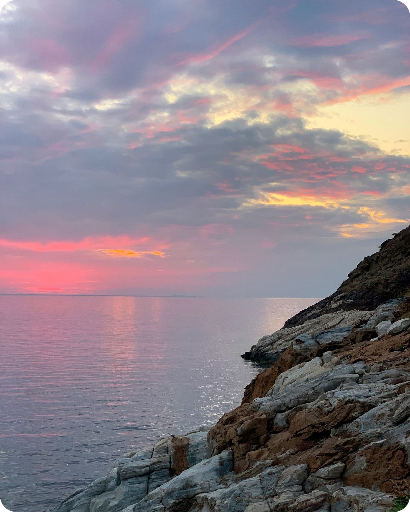 Rocky shoreline at sunset with a colorful sky reflecting on calm water, featuring pink, purple, and orange hues under a cloudy sky.