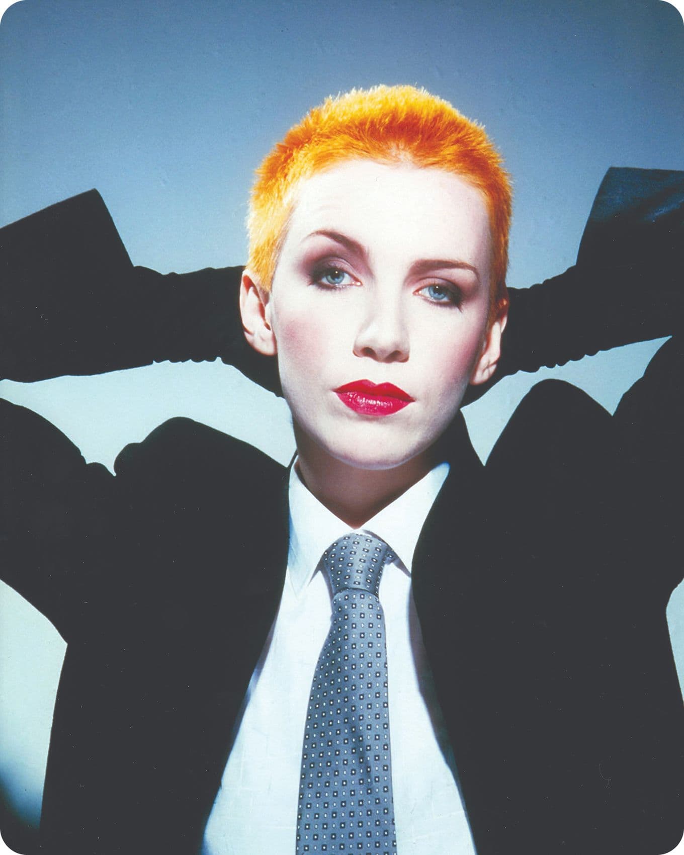 Annie Lennox with short orange hair, wearing a suit and tie, poses with hands behind head against a blue-gray background.