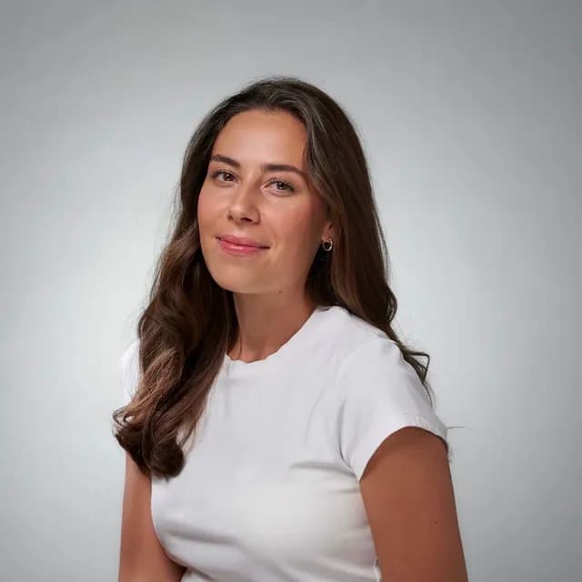 Woman with long brown hair smiling slightly, wearing a white t-shirt, against a plain gray background.