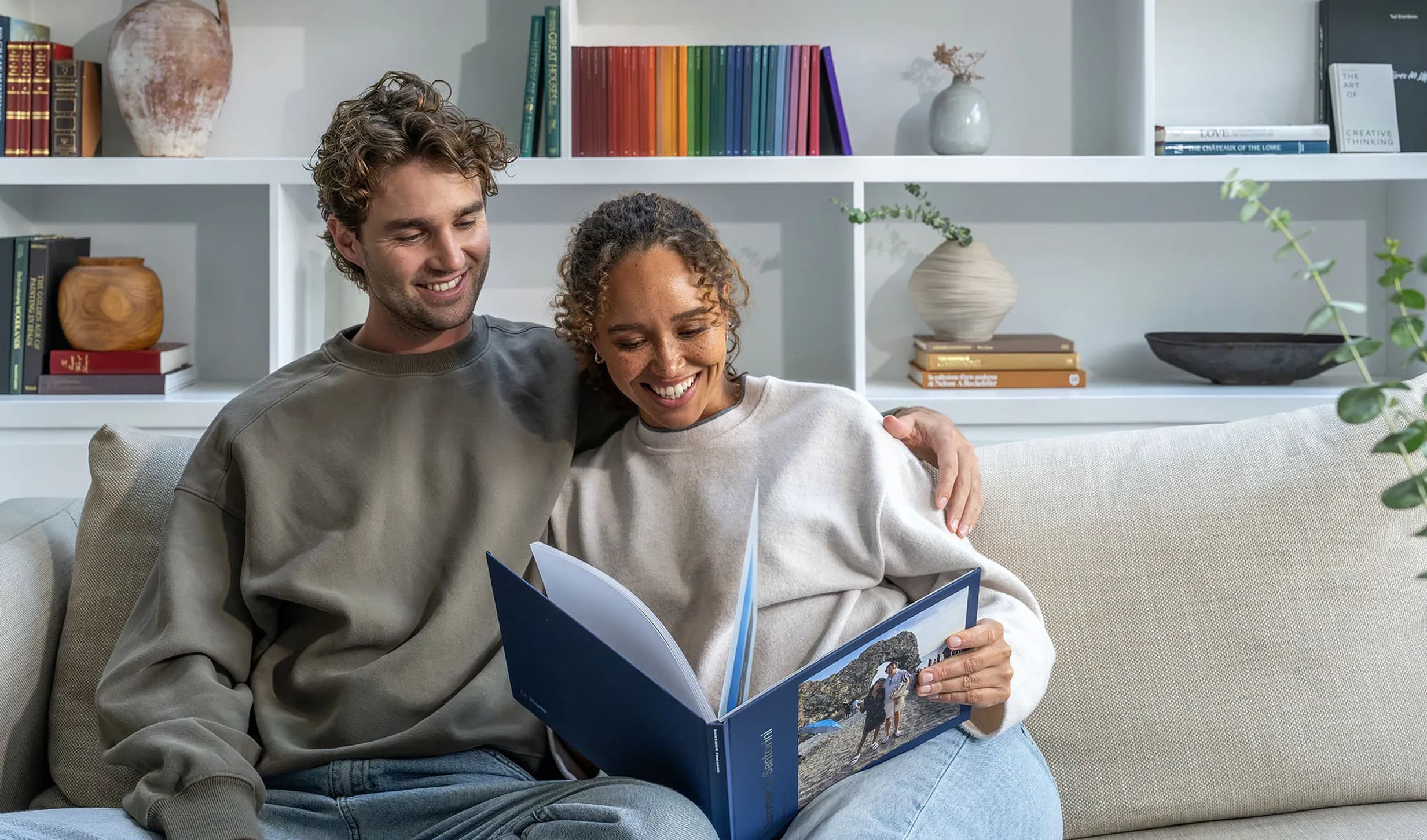 A smiling couple on a sofa looks at a Popsa photo album, open to a picture of a rocky beach. White shelves with books and decor are in the background.