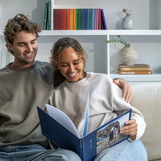 A smiling couple on a sofa looks at a Popsa photo album, open to a picture of a rocky beach. White shelves with books and decor are in the background.