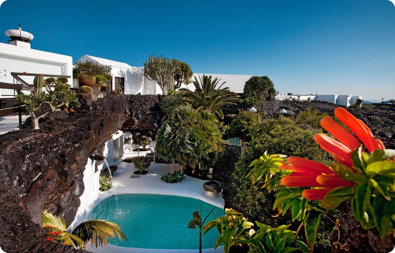 A tropical garden with lush greenery surrounds a turquoise pool, set against a backdrop of white buildings and a clear blue sky.