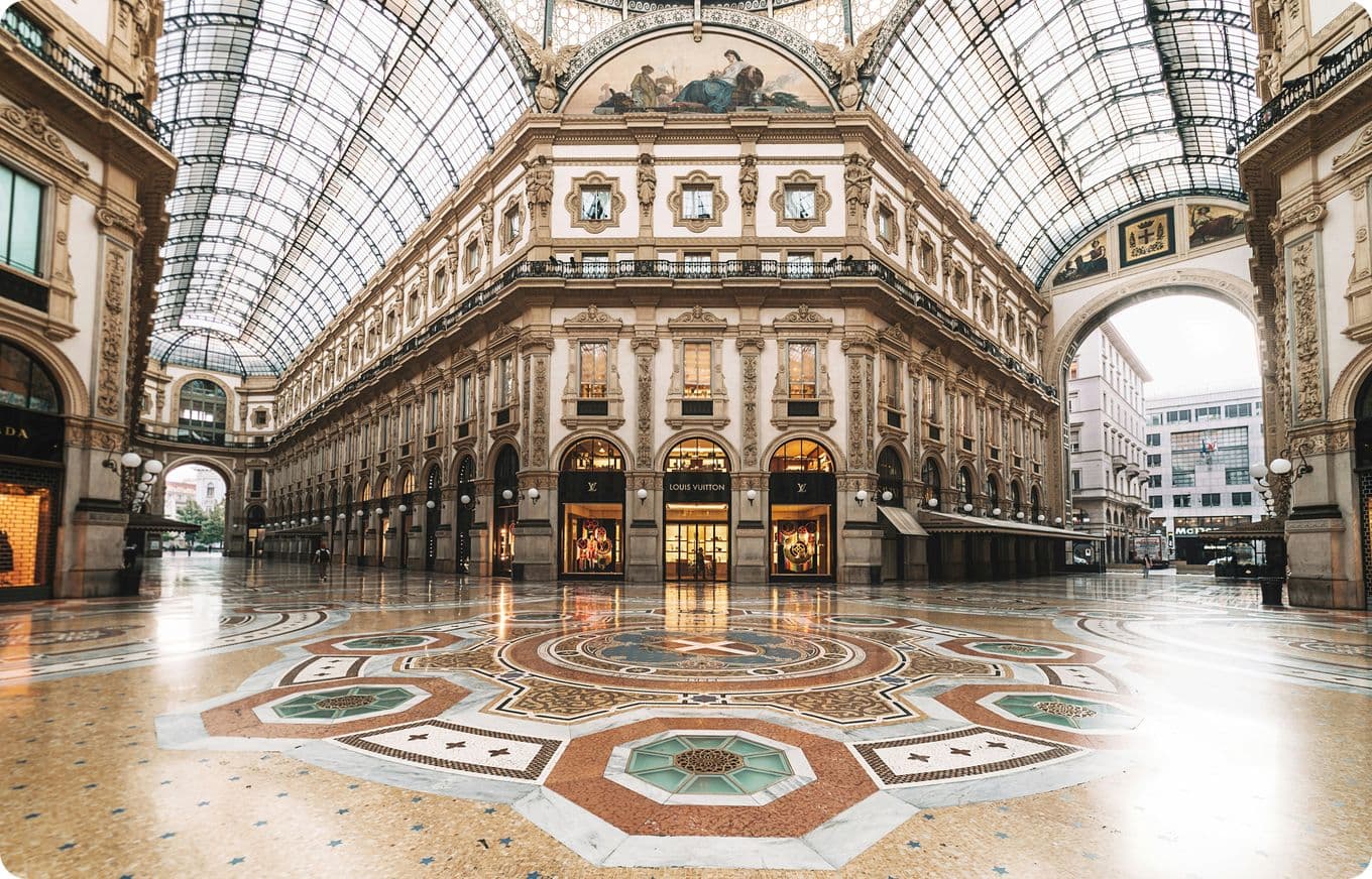 Ornate, empty shopping arcade with a glass ceiling, intricate floor mosaic, and elegant architecture featuring arches and detailed facades.