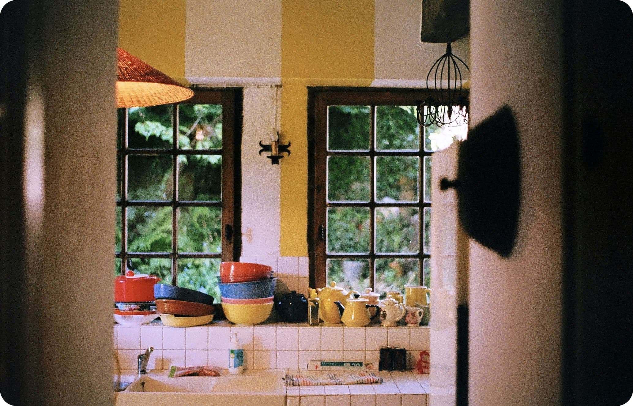 Cozy kitchen with yellow walls, stacked colorful dishes, and large windows overlooking lush greenery.