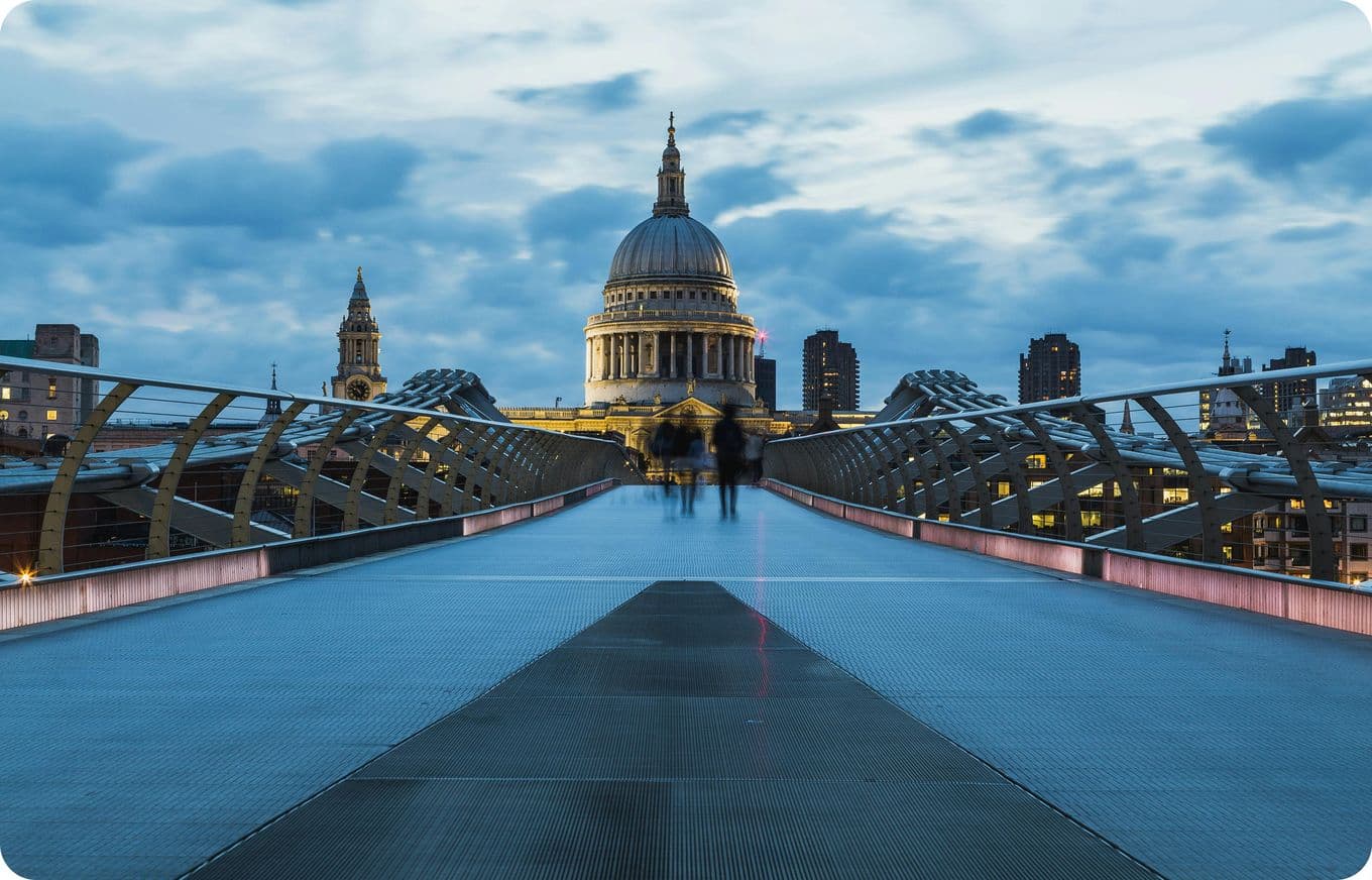 View of St. Paul's Cathedral from the Millennium Bridge in London at dusk, with a cloudy sky and blurred pedestrians on the bridge.