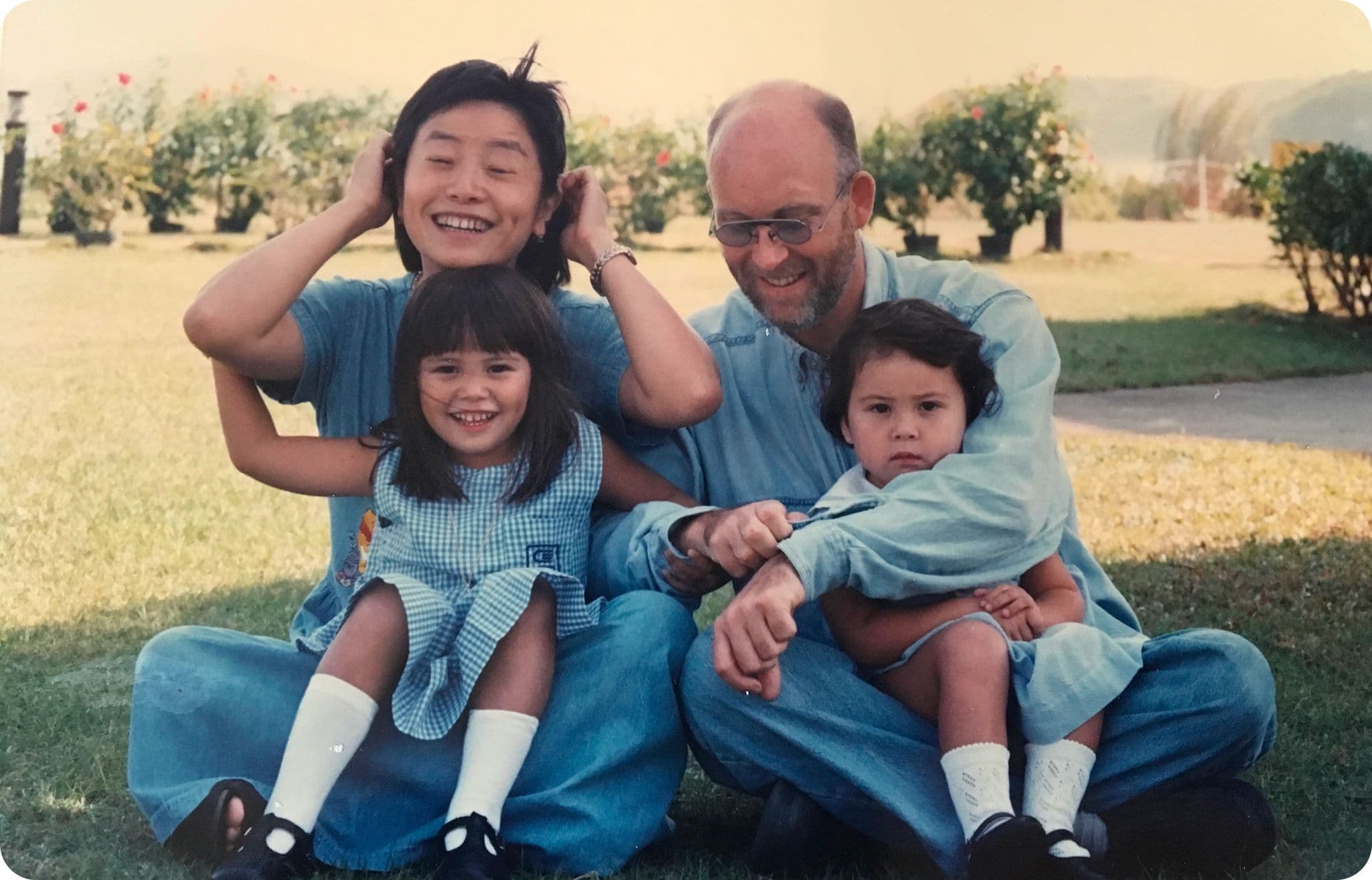 A family of four sitting on grass; two adults and two children smiling, dressed casually, with bushes and mountains in the background.