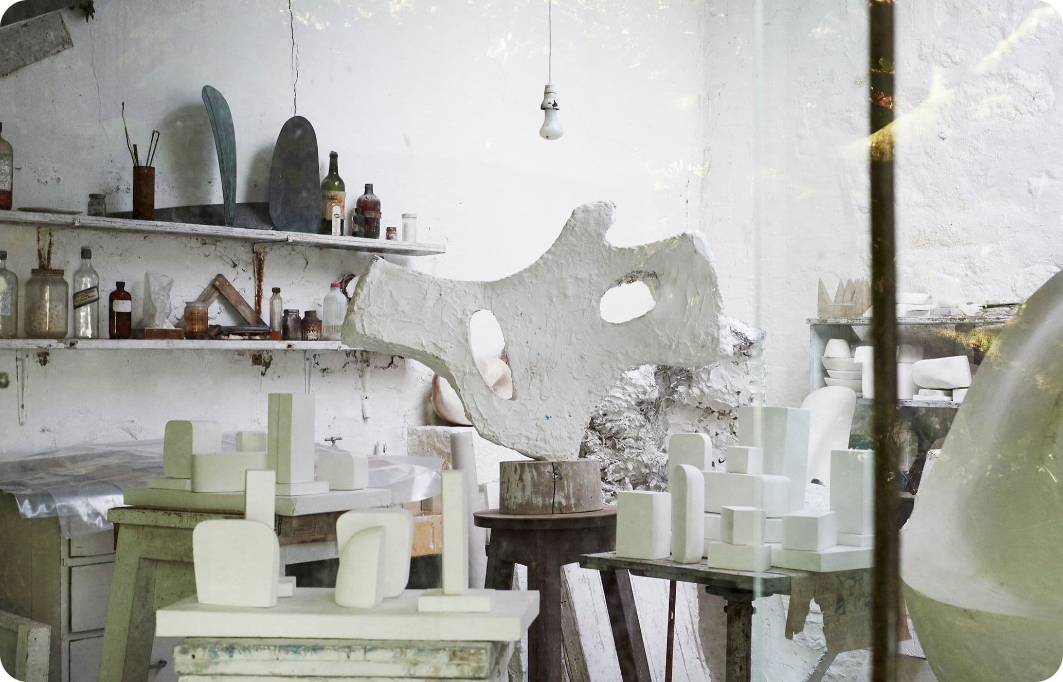 Artist's studio with abstract sculptures, shelves with bottles, and a hanging light bulb. The space is filled with white and gray tones.