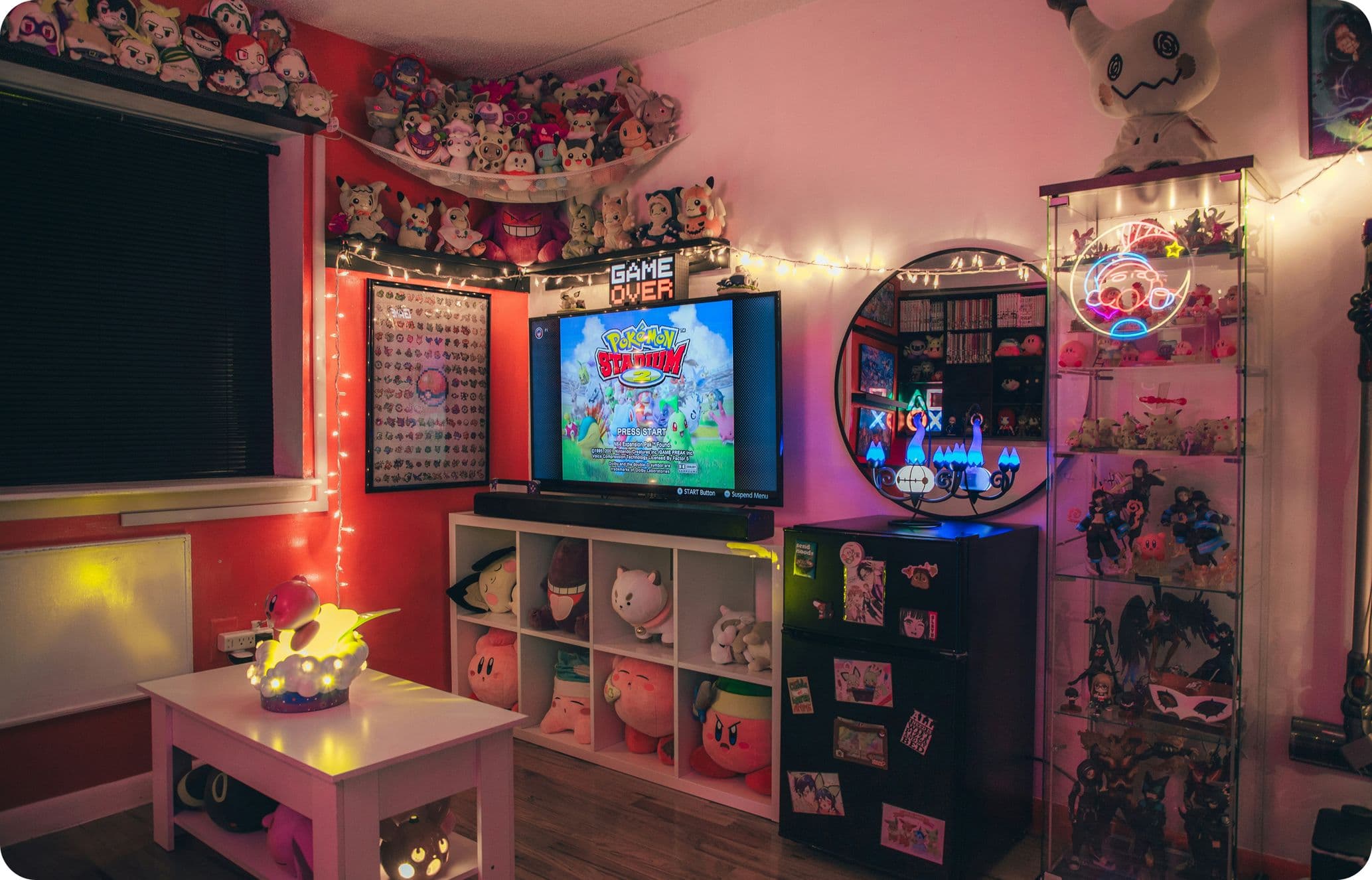 Cozy gaming room with a TV displaying a colorful game, surrounded by plush toys, shelves, neon lights, and gaming memorabilia.