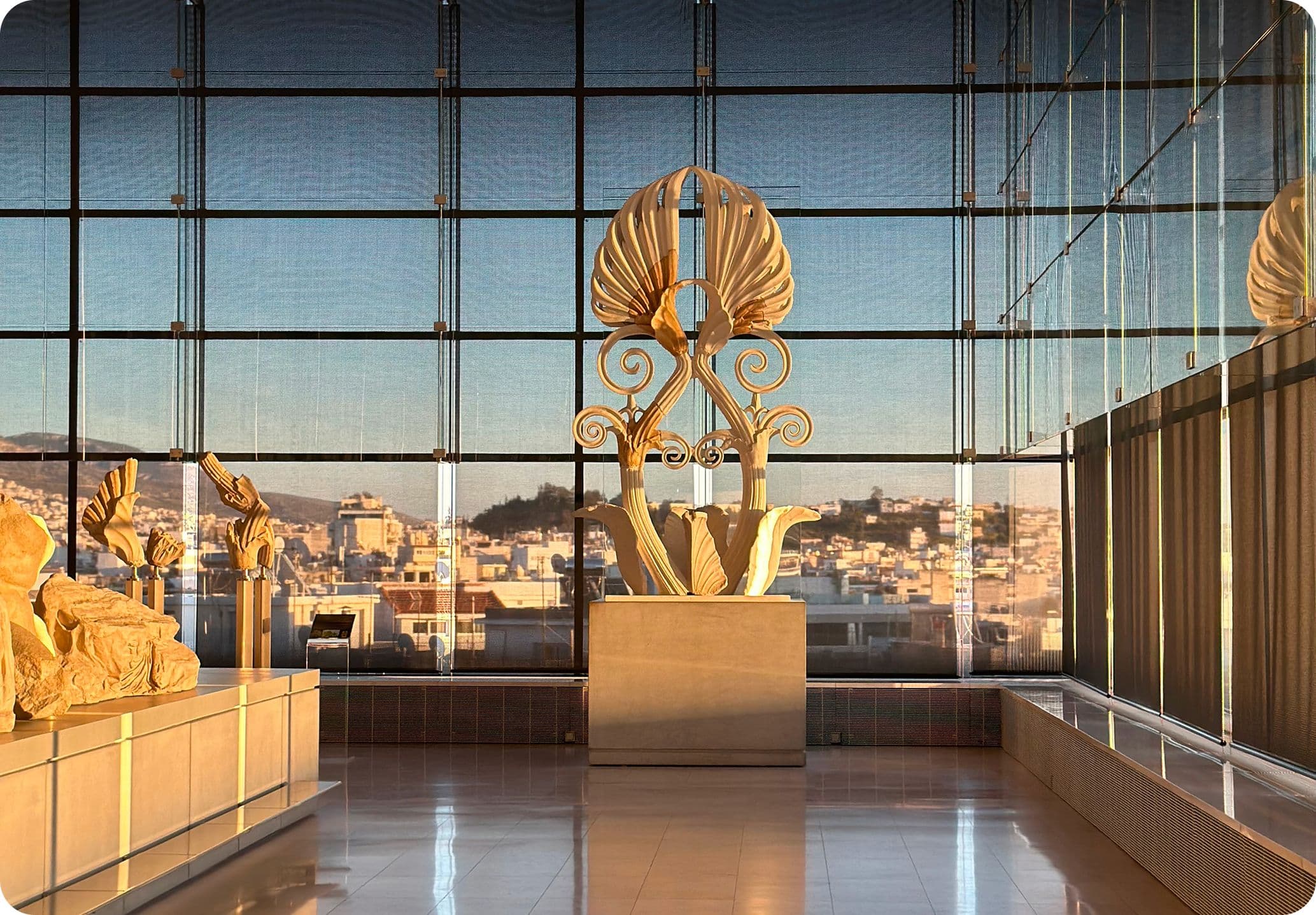 Museum interior with large windows showing a city view. Ancient Greek sculptures and artifacts are displayed in a sunlit gallery.