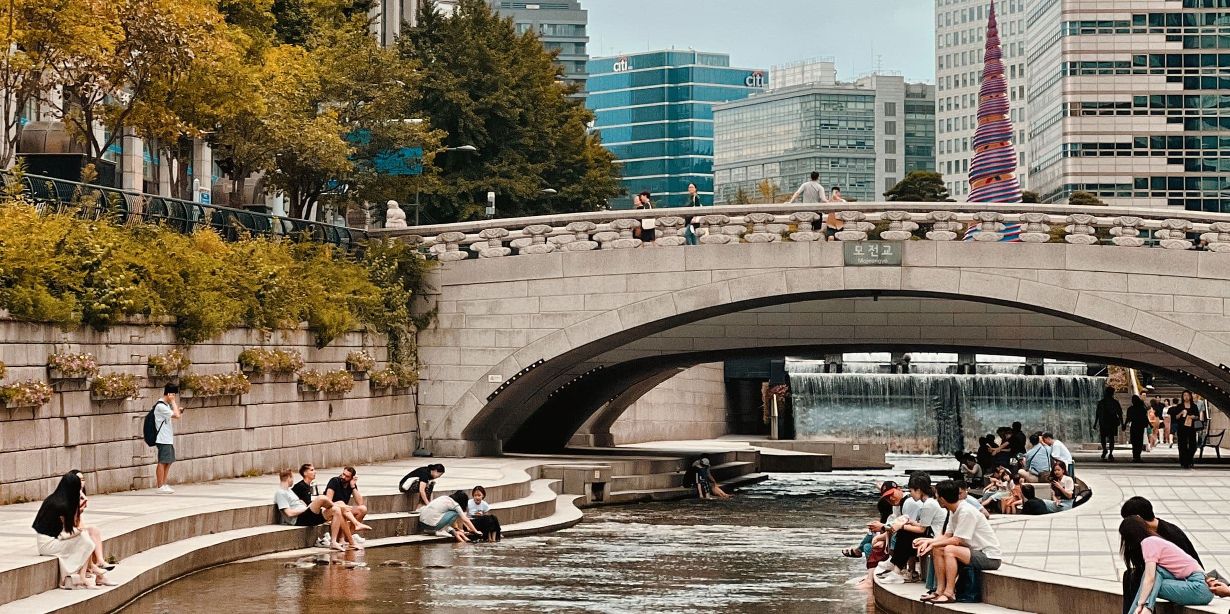 People relax by a riverside under a stone bridge in an urban park, surrounded by trees and modern buildings.