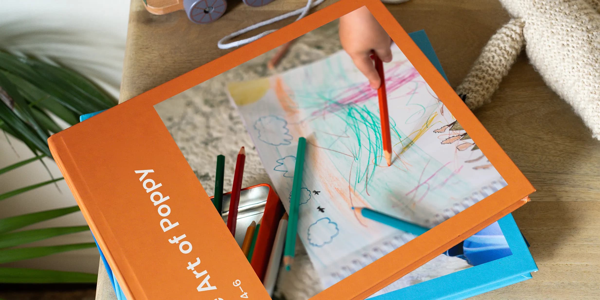 A child's hand draws with colored pencils on paper next to an orange book titled "Art of Poppy" on a wooden table.