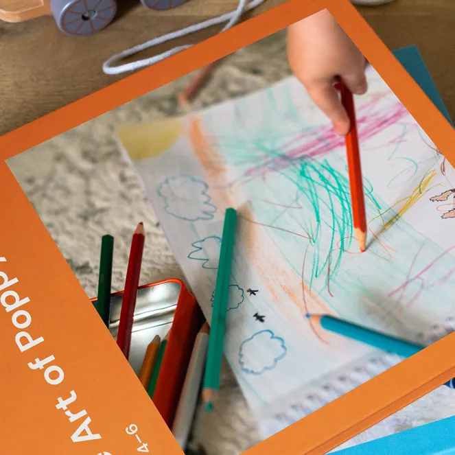 A child's hand draws with colored pencils on paper next to an orange book titled "Art of Poppy" on a wooden table.