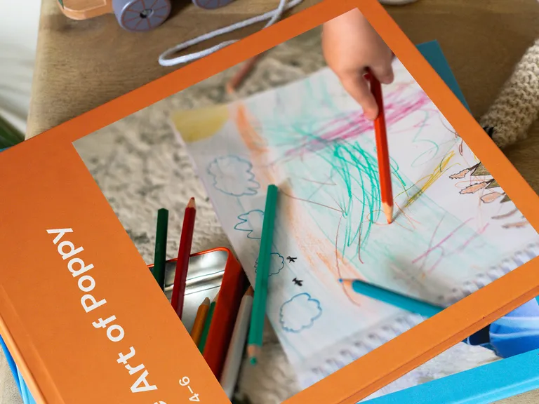 A child's hand draws with colored pencils on paper next to an orange book titled "Art of Poppy" on a wooden table.