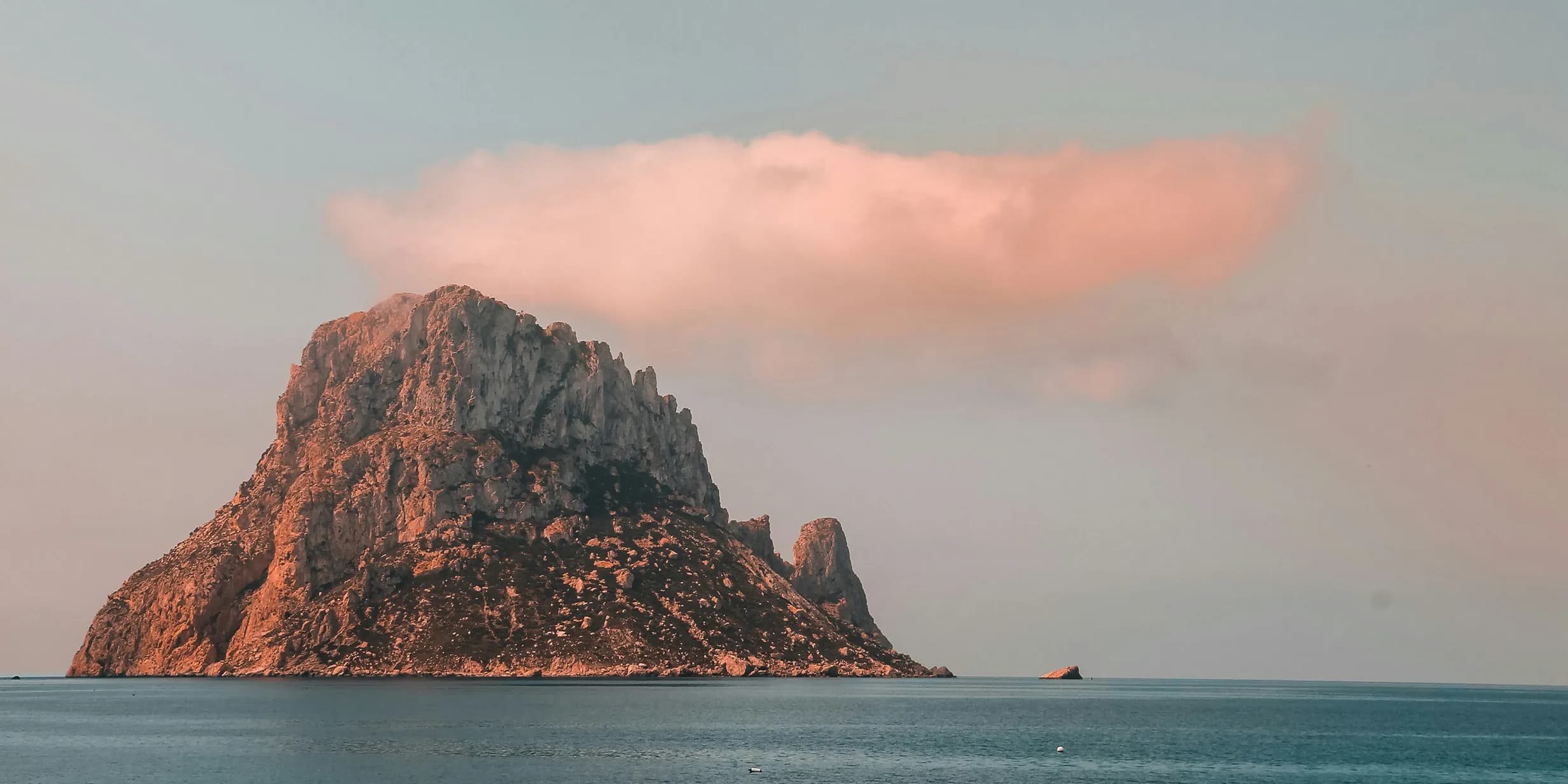 Rocky island rising from the sea under a pinkish cloud during sunset.