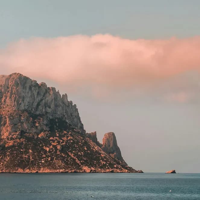 Rocky island rising from the sea under a pinkish cloud during sunset.