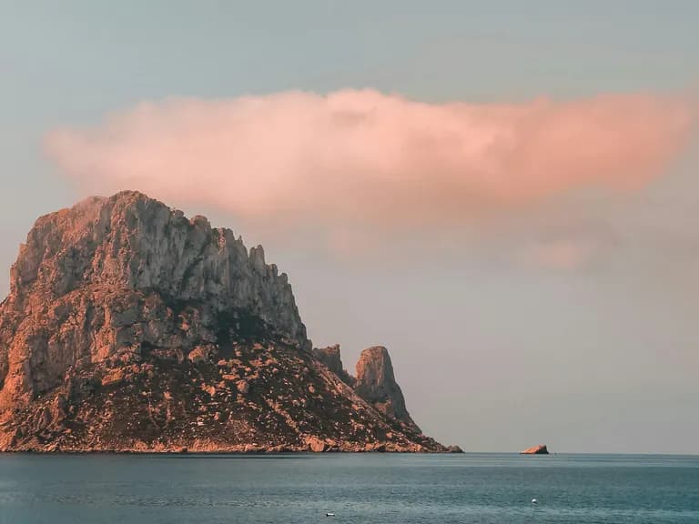 Rocky island rising from the sea under a pinkish cloud during sunset.