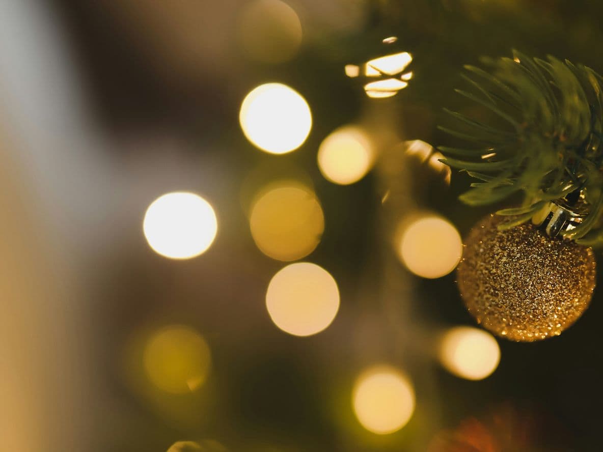 Close-up of a Christmas tree with a gold ornament. Blurred golden lights sparkle softly in the background.