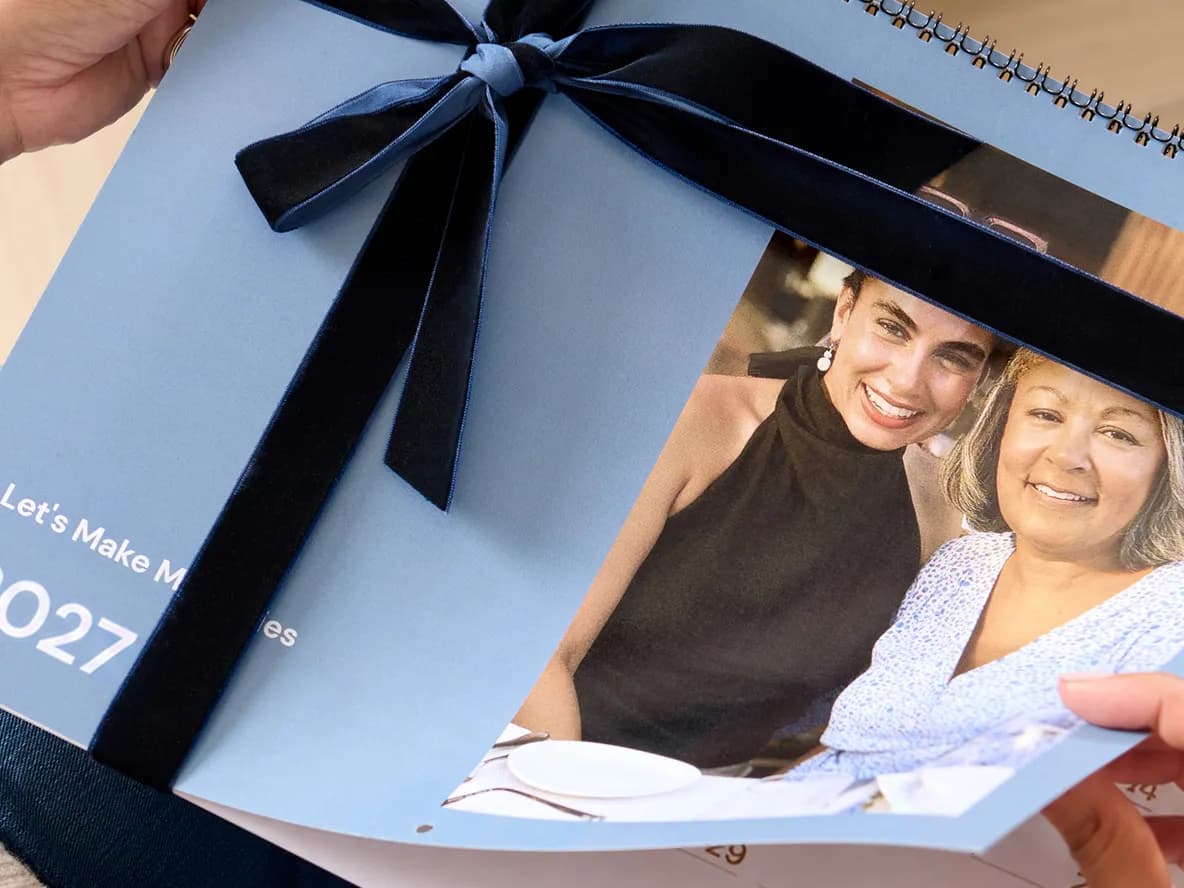 A person holding a 2027 spiral-bound photo calendar with a blue cover, adorned with a black ribbon and featuring a photo of two smiling women.