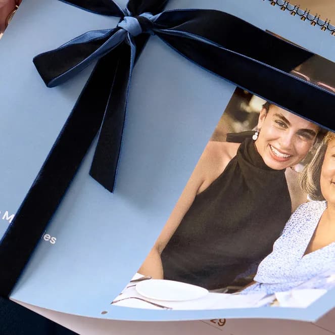 A person holding a 2027 spiral-bound photo calendar with a blue cover, adorned with a black ribbon and featuring a photo of two smiling women.