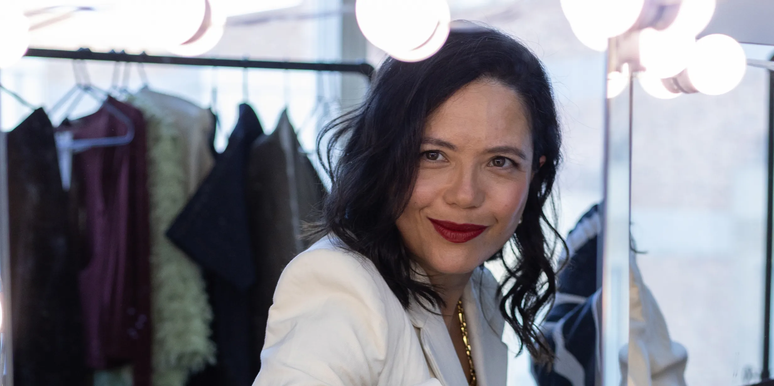 A woman with dark hair and red lipstick smiles at the camera in a well-lit dressing room, with clothes hanging in the background.
