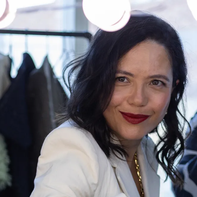 A woman with dark hair and red lipstick smiles at the camera in a well-lit dressing room, with clothes hanging in the background.