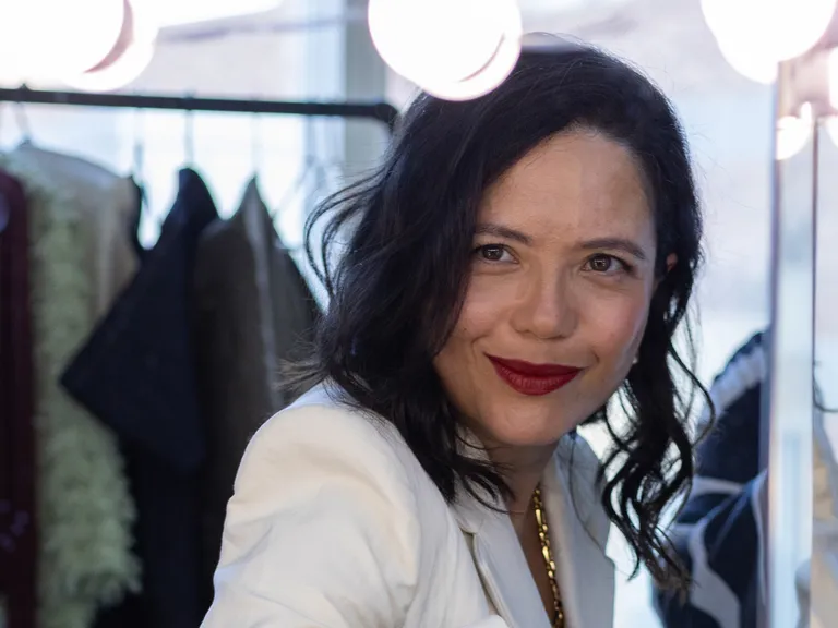 A woman with dark hair and red lipstick smiles at the camera in a well-lit dressing room, with clothes hanging in the background.