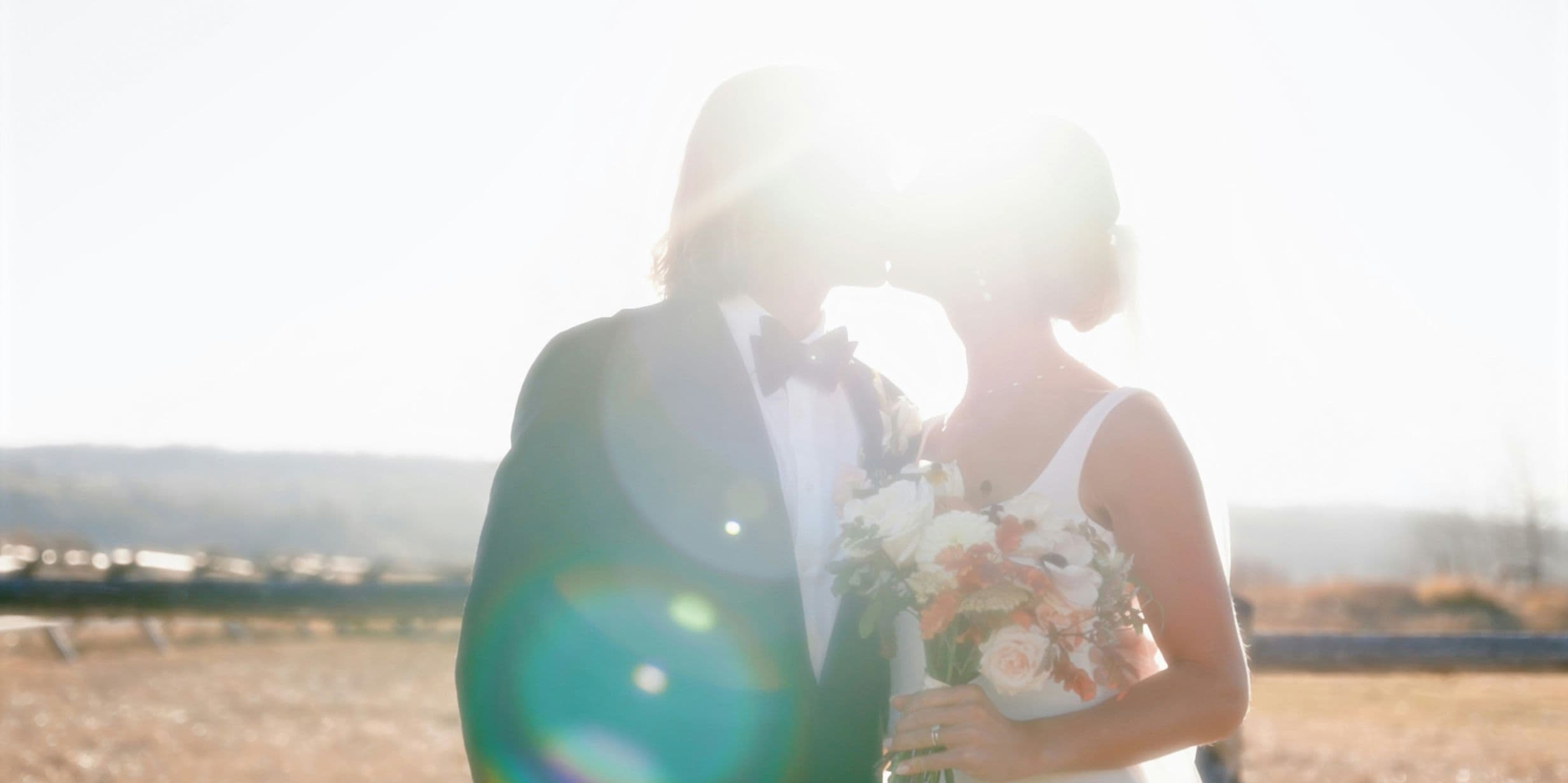 A couple in wedding attire kiss in bright sunlight, holding a bouquet, with lens flare and a blurred natural background.