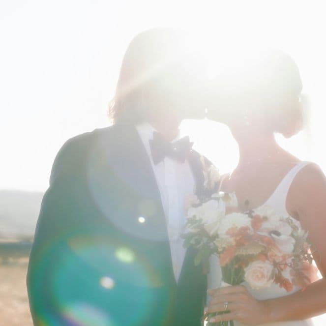A couple in wedding attire kiss in bright sunlight, holding a bouquet, with lens flare and a blurred natural background.
