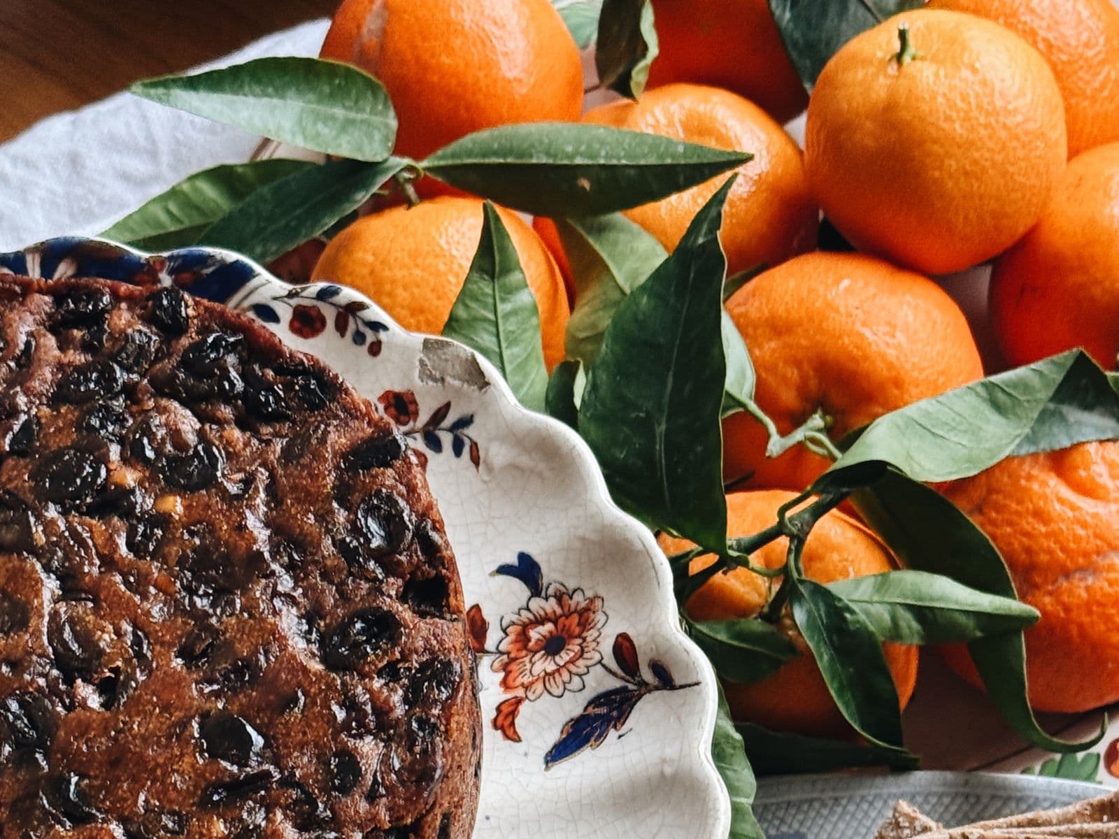 Fruitcake on a floral plate next to a bowl of fresh oranges with green leaves on a wooden table.