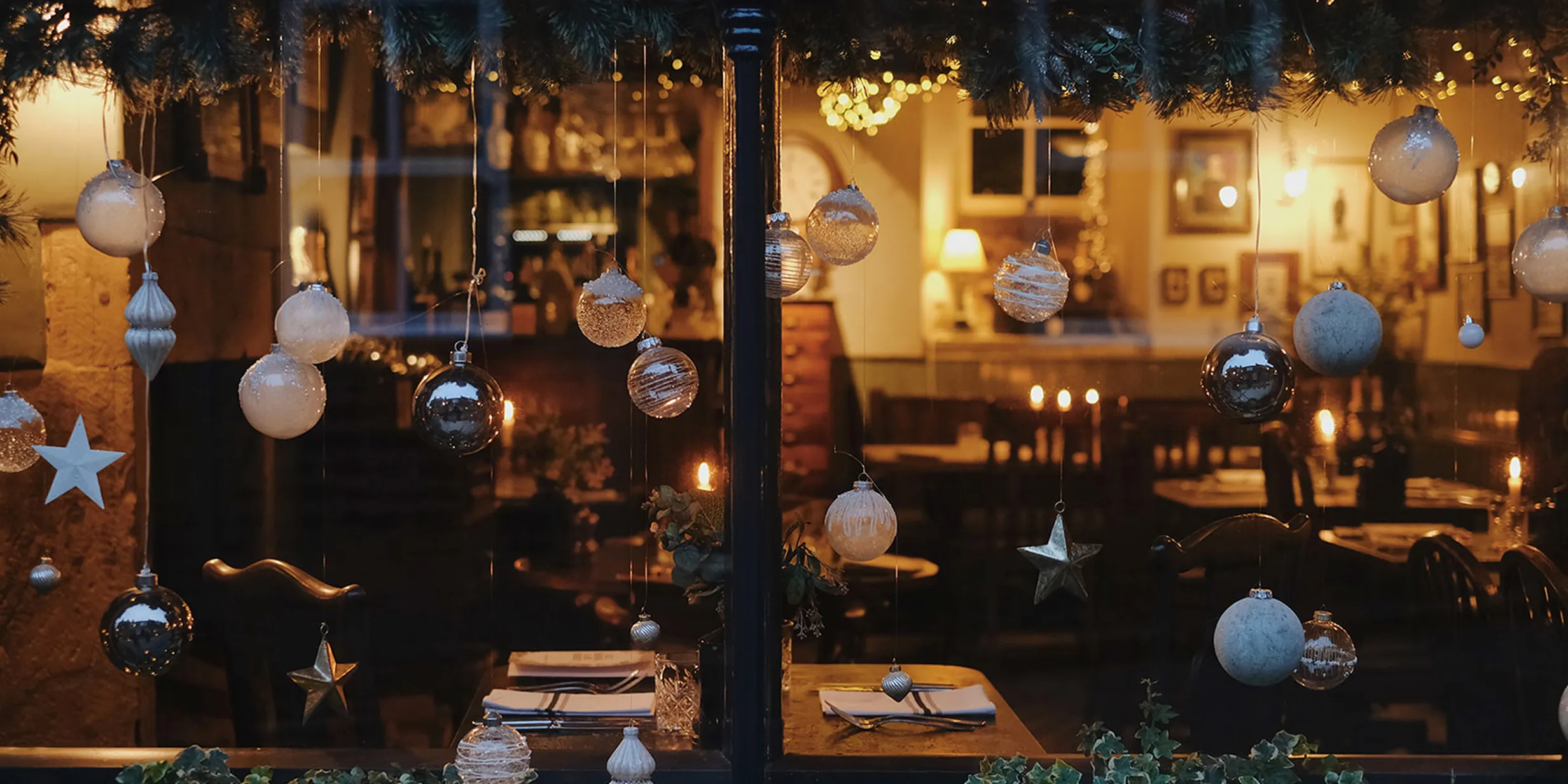 Cozy restaurant interior viewed through a window decorated with hanging ornaments and lights, tables set for dinner, and warm ambient lighting.