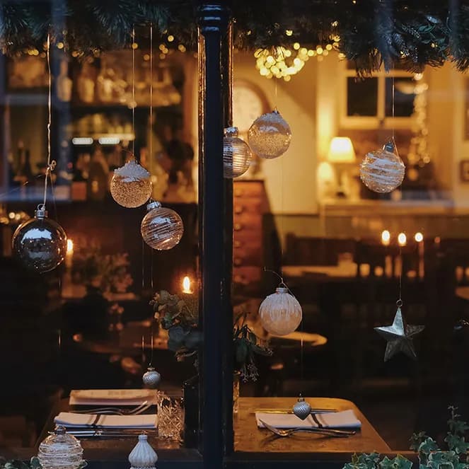 Cozy restaurant interior viewed through a window decorated with hanging ornaments and lights, tables set for dinner, and warm ambient lighting.